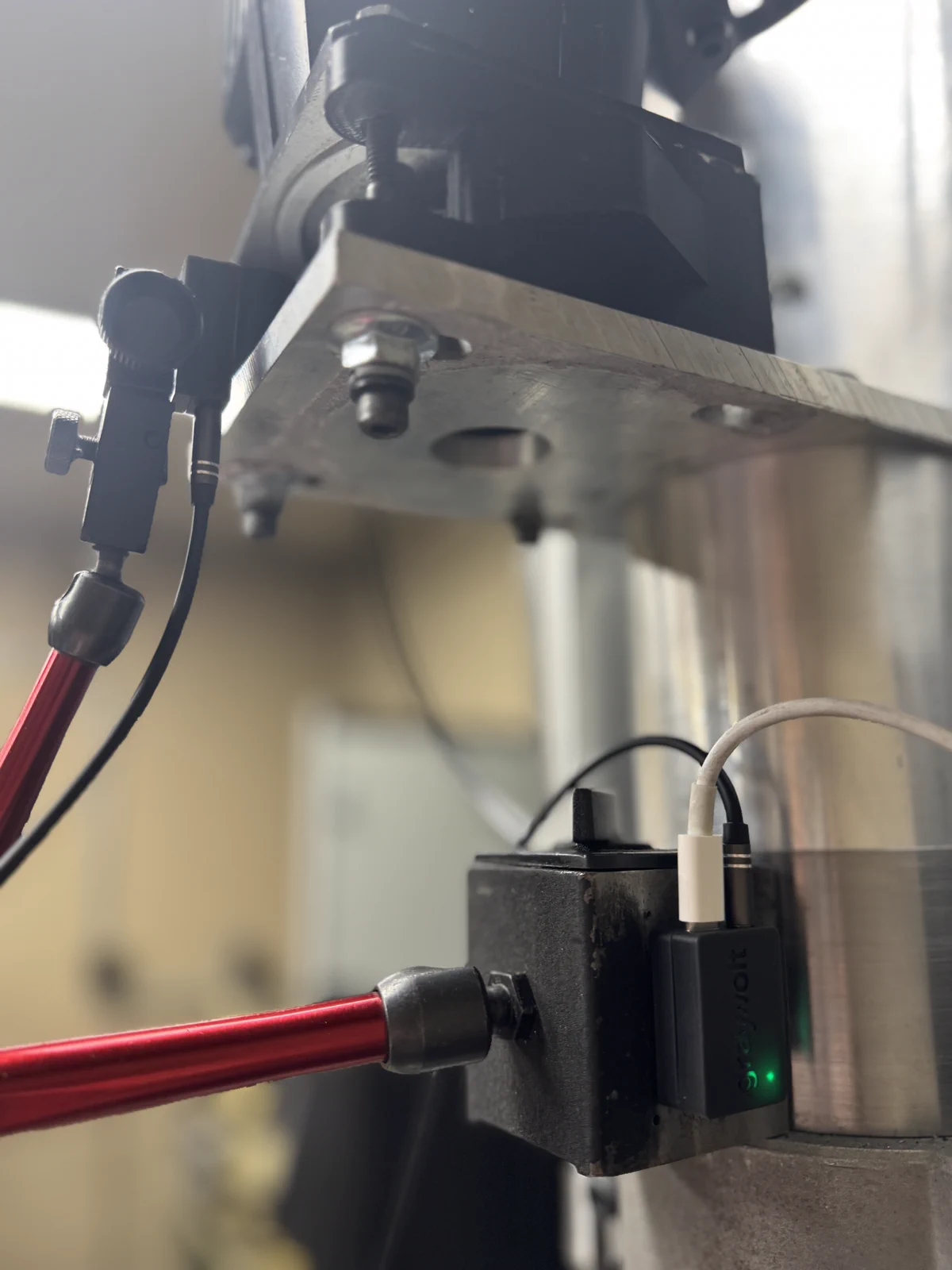 GrayVolt unit magnetically mounted on a CNC spindle with the wired IR tach pickup on a red dial indicator stand, a black 3.5 millimeter tach cable, and a white USB-C power cable connected